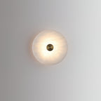 Front view of the lit alabaster disc sconce, the brass cap set flush in the pale stone face.