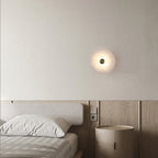 Alabaster disc wall light lit above a wooden nightstand beside a bed with striped bedding.