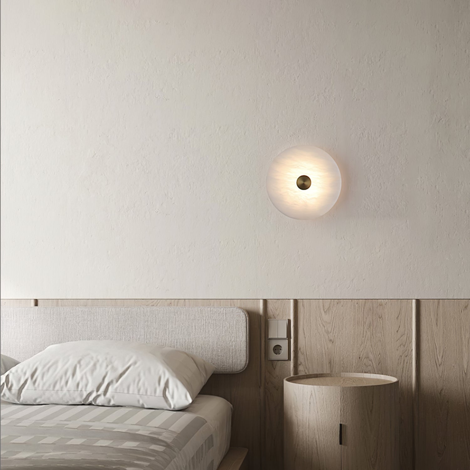 Alabaster disc wall light lit above a wooden nightstand beside a bed with striped bedding.