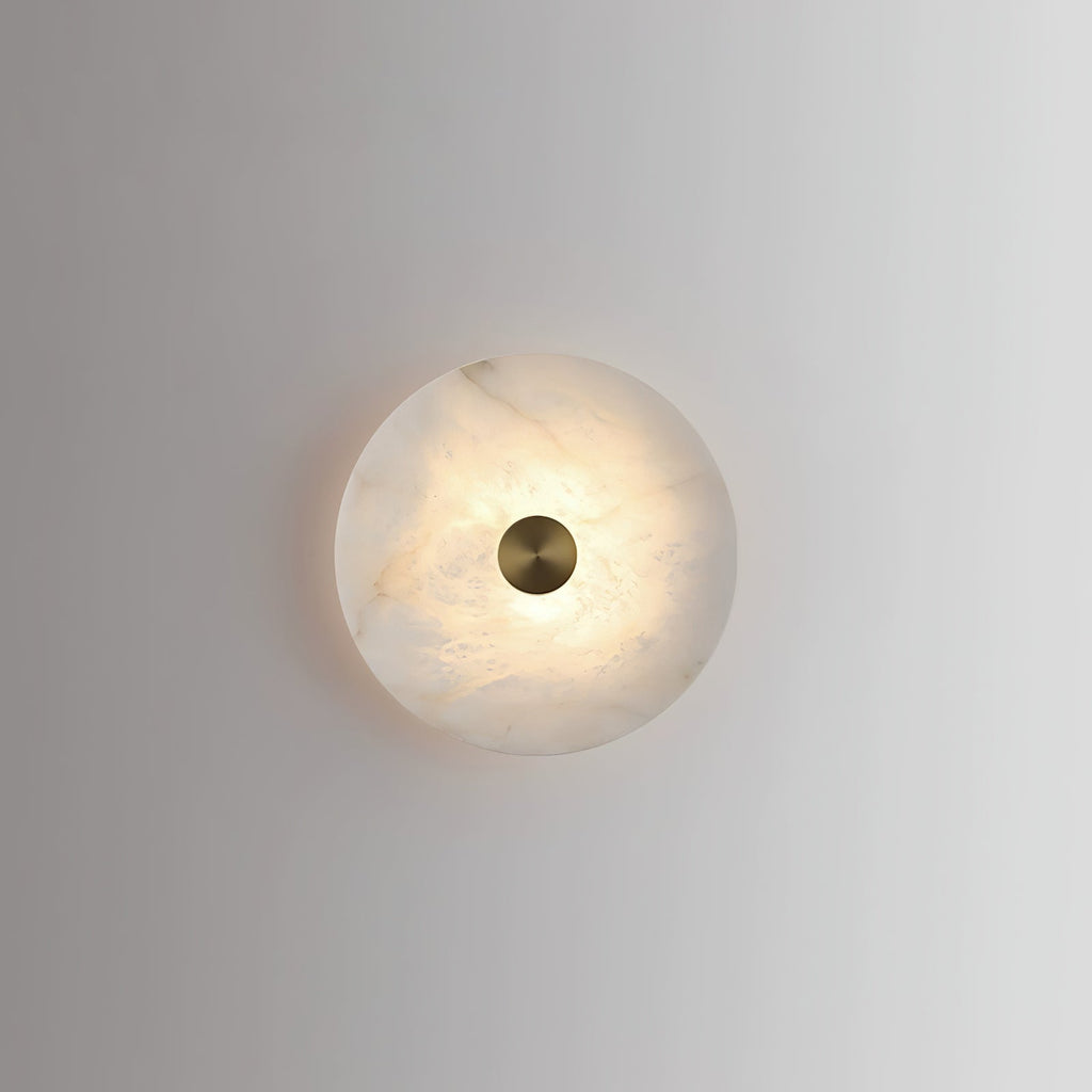 Single alabaster disc wall light lit on a plain grey wall, casting a soft halo around itself.