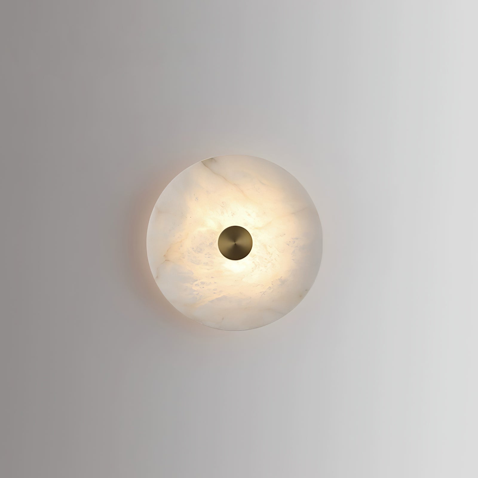 Single alabaster disc wall light lit on a plain grey wall, casting a soft halo around itself.
