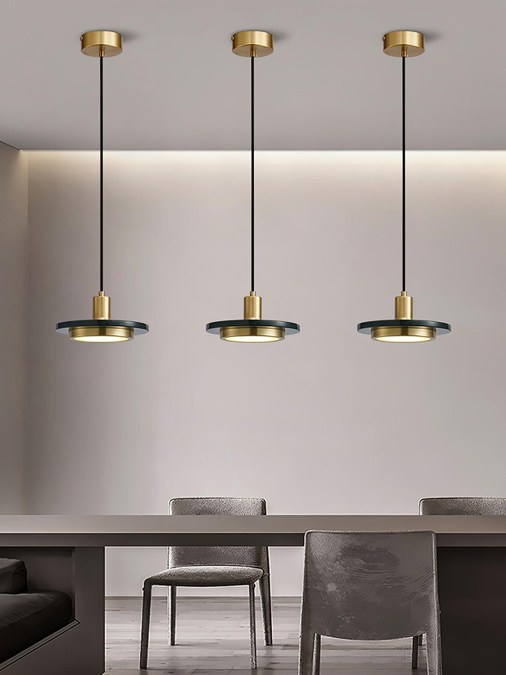 Three gold Clérachic pendant lights spaced along a wall above a dining table in a minimalist grey room.