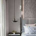 A black Clérachic pendant light hangs beside a grey upholstered bed with a potted plant on the nightstand.
