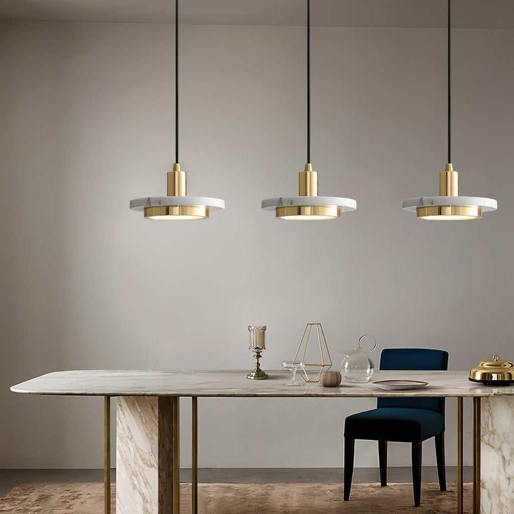 Three gold Clérachic pendant lights hang above a marble dining table with gold legs and a blue velvet chair.