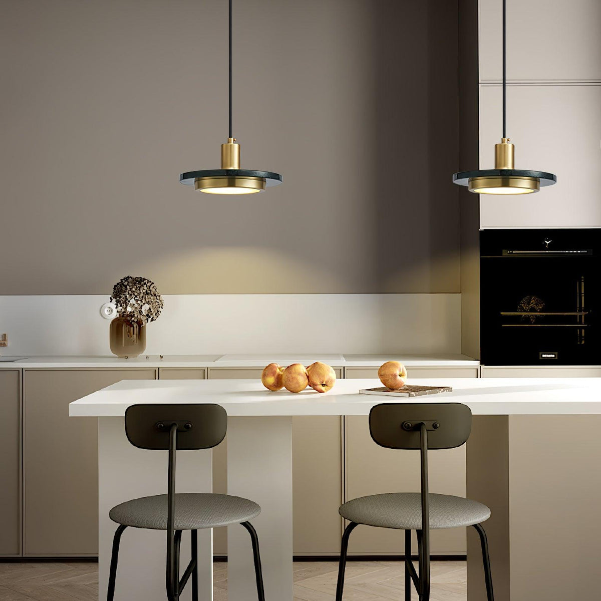 Two gold Clérachic pendant lights hang above a beige kitchen island lined with stools and a bowl of fruit.