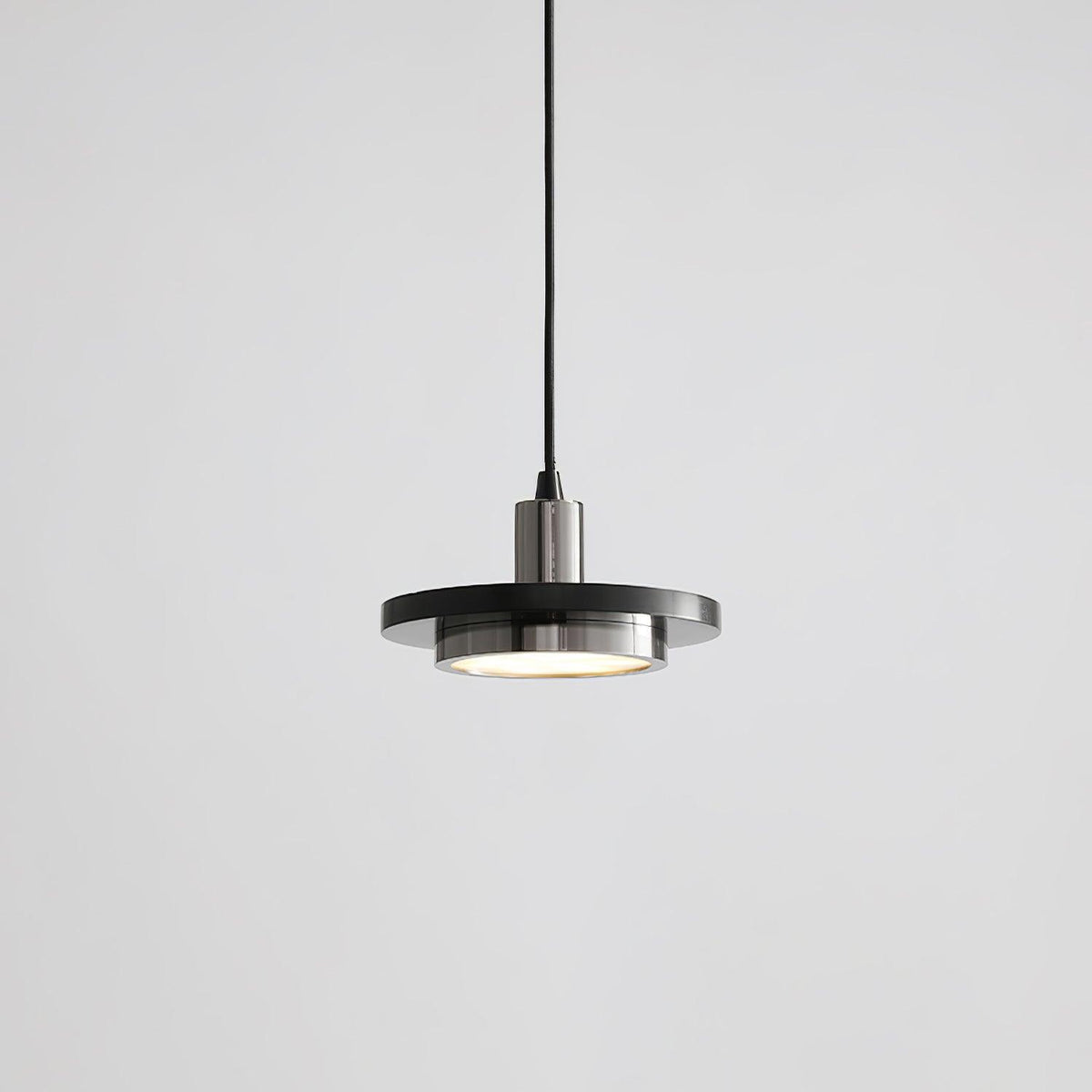 A matt dark disc pendant with a brushed steel-toned fitting drops on its black cord in this grey-background shot from Clérachic.