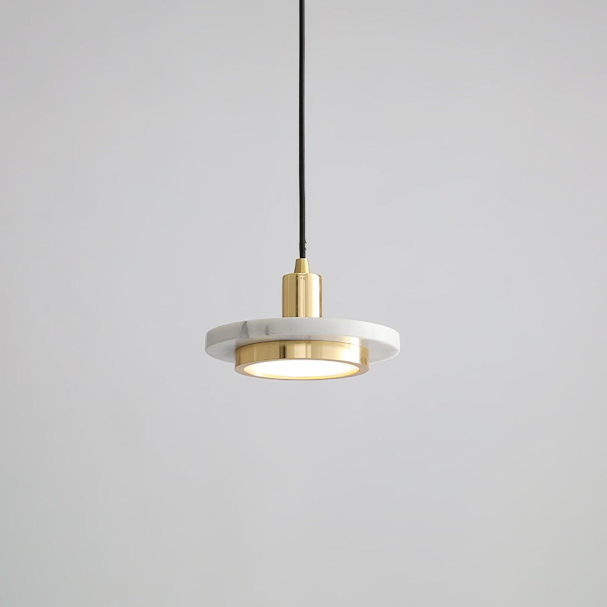 White marble disc pendant with a gold-toned rim caught mid-cord against grey, the Clérachic fixture's brass fitting bright at its centre.