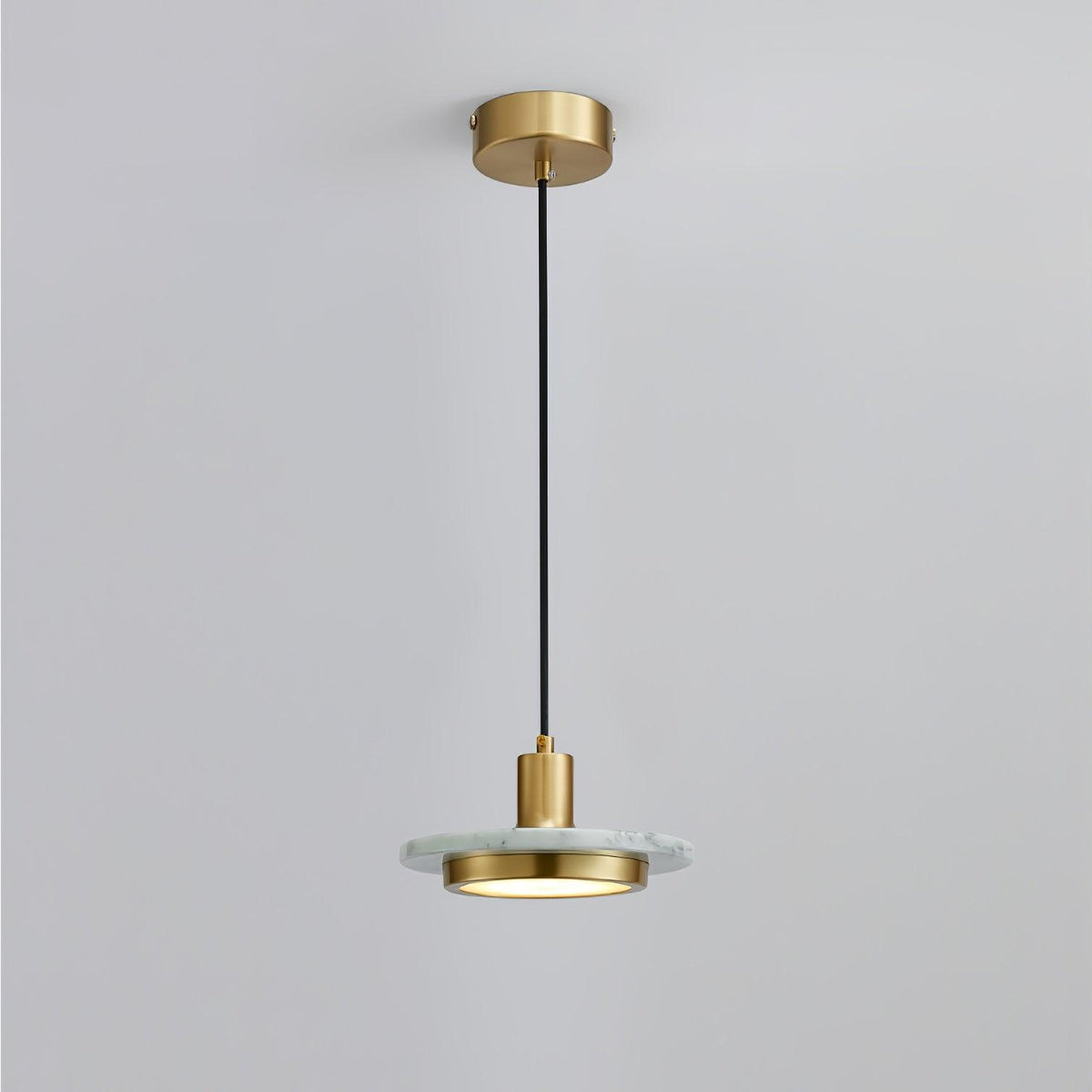 A pale marble disc pendant hangs on a long black cord from its brass ceiling plate, photographed full length by Clérachic against soft grey.