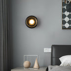 Wider view of the same Clérachic black marble wall sconce lit in the bedroom, showing more of the grey wall and headboard.