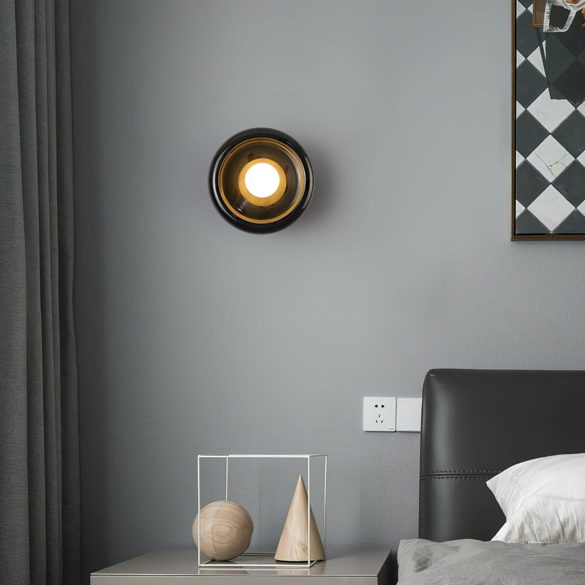 Wider view of the same Clérachic black marble wall sconce lit in the bedroom, showing more of the grey wall and headboard.