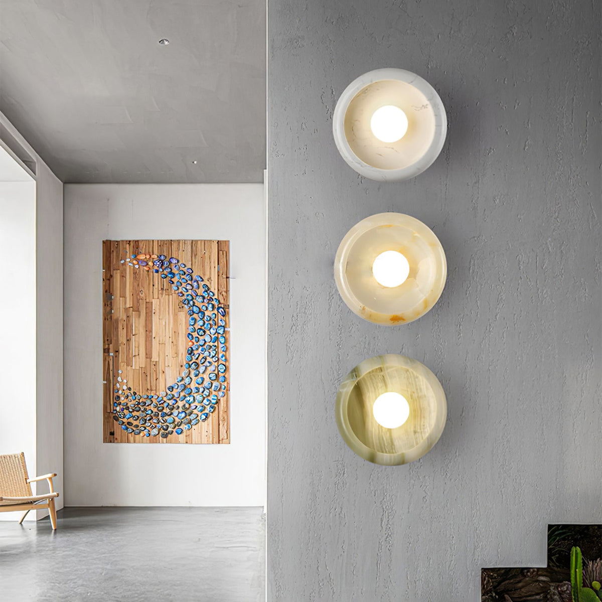 Three Clérachic jade wall sconces in different stone tones lit along a hallway wall beside a wooden mosaic art panel.