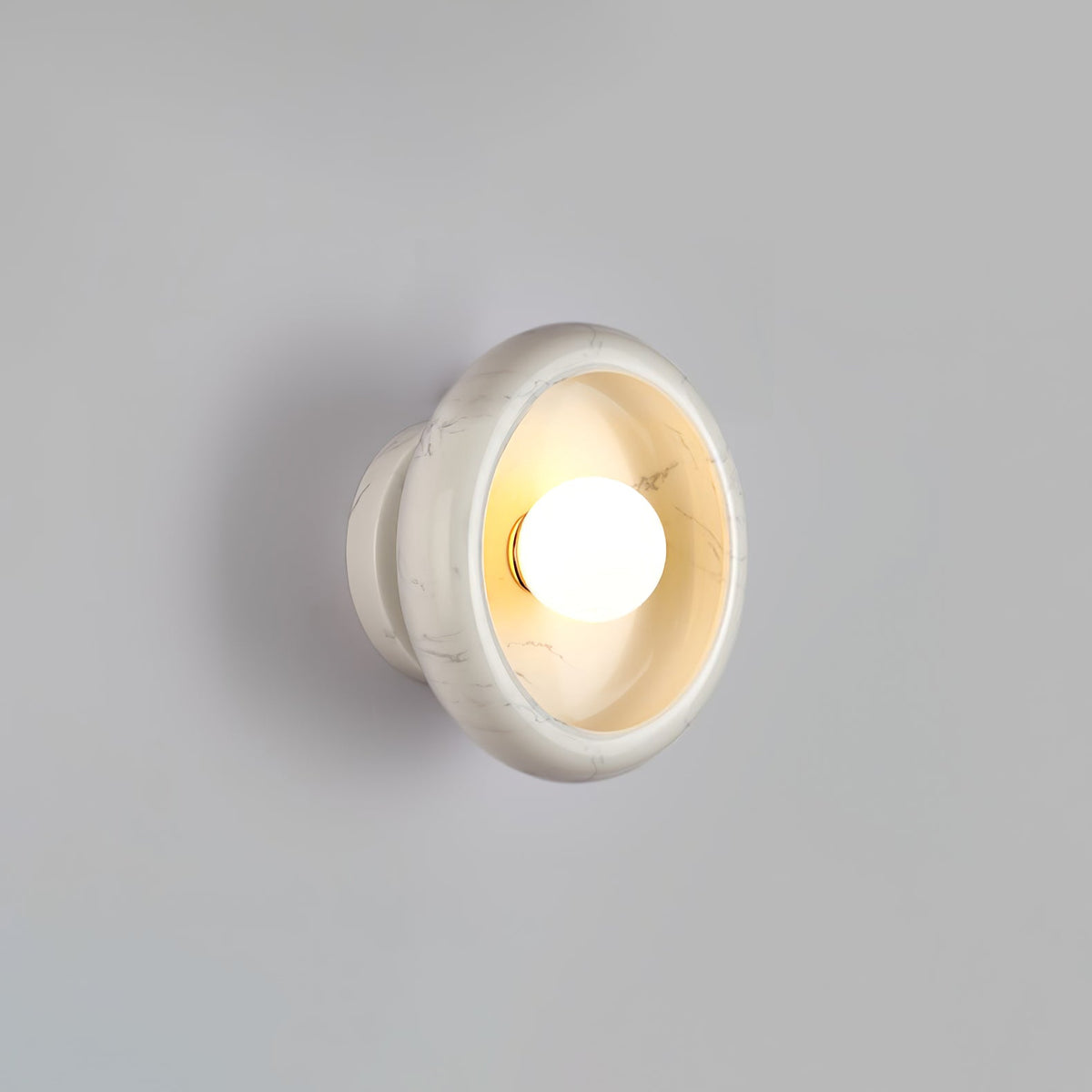 Close-up of a Clérachic white marble wall sconce lit against a grey wall, its veined interior visible around the bulb.