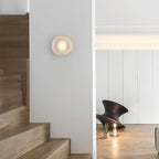 Clérachic white wall sconce lit in a stairwell hallway, glowing beside wooden stairs and a small sculptural object.
