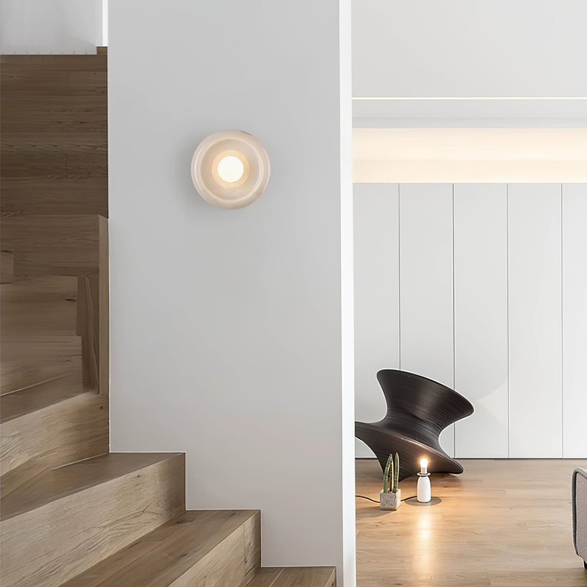 Clérachic white wall sconce lit in a stairwell hallway, glowing beside wooden stairs and a small sculptural object.