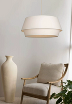 The white linen shade of this Clérachic pendant hangs beside a tall cream vase and a wood-framed armchair.
