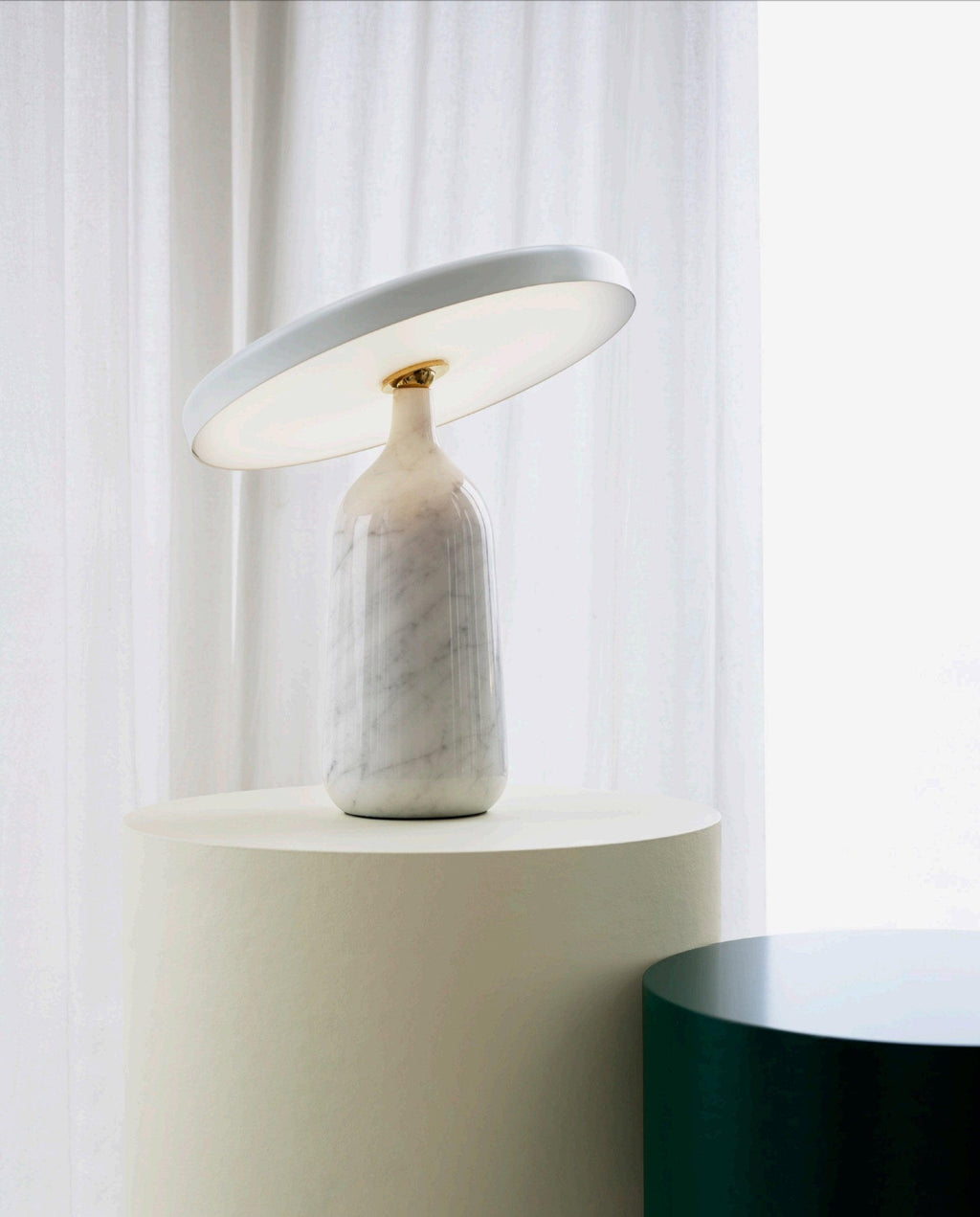 Clérachic's white marble table lamp sits on a round pedestal beside a sheer curtain, its disc shade angled toward the window light.