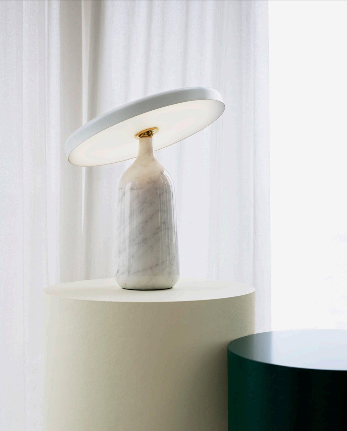 Clérachic's white marble table lamp sits on a round pedestal beside a sheer curtain, its disc shade angled toward the window light.