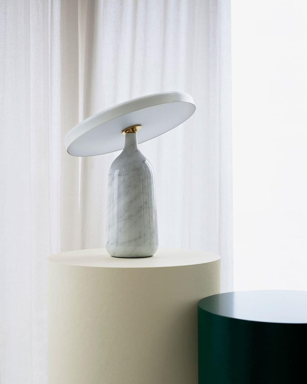 White marble Clérachic table lamp on a round cream pedestal beside a sheer curtain, its disc shade tilted upward.
