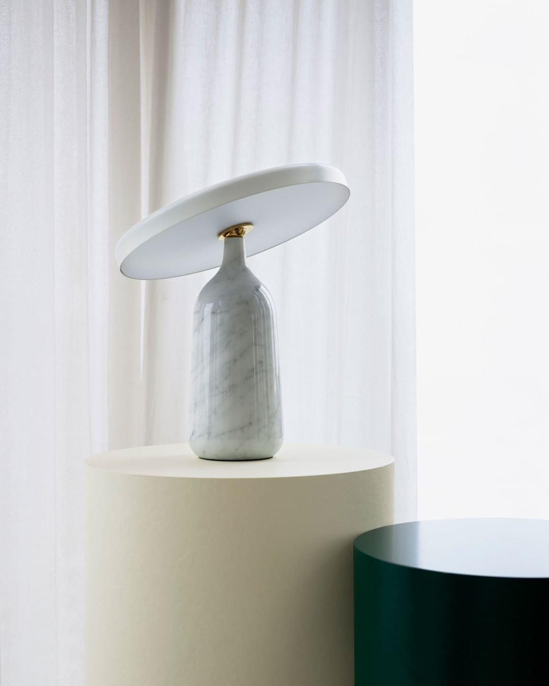 White marble Clérachic table lamp on a round cream pedestal beside a sheer curtain, its disc shade tilted upward.