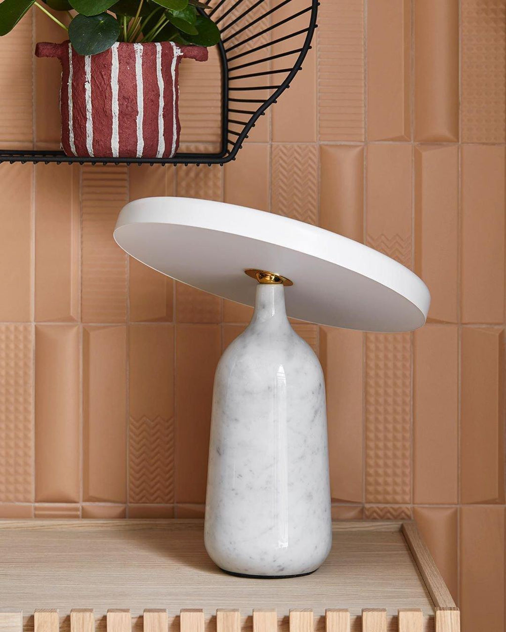 White marble Clérachic table lamp on a wood-slat shelf against a terracotta patterned tile wall, shade angled to one side.
