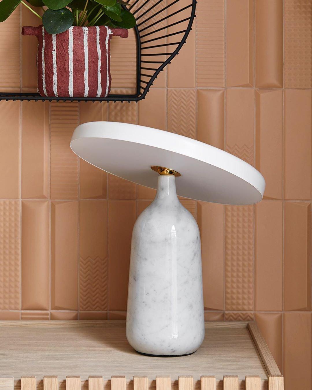 White marble Clérachic table lamp on a wood-slat shelf against a terracotta patterned tile wall, shade angled to one side.
