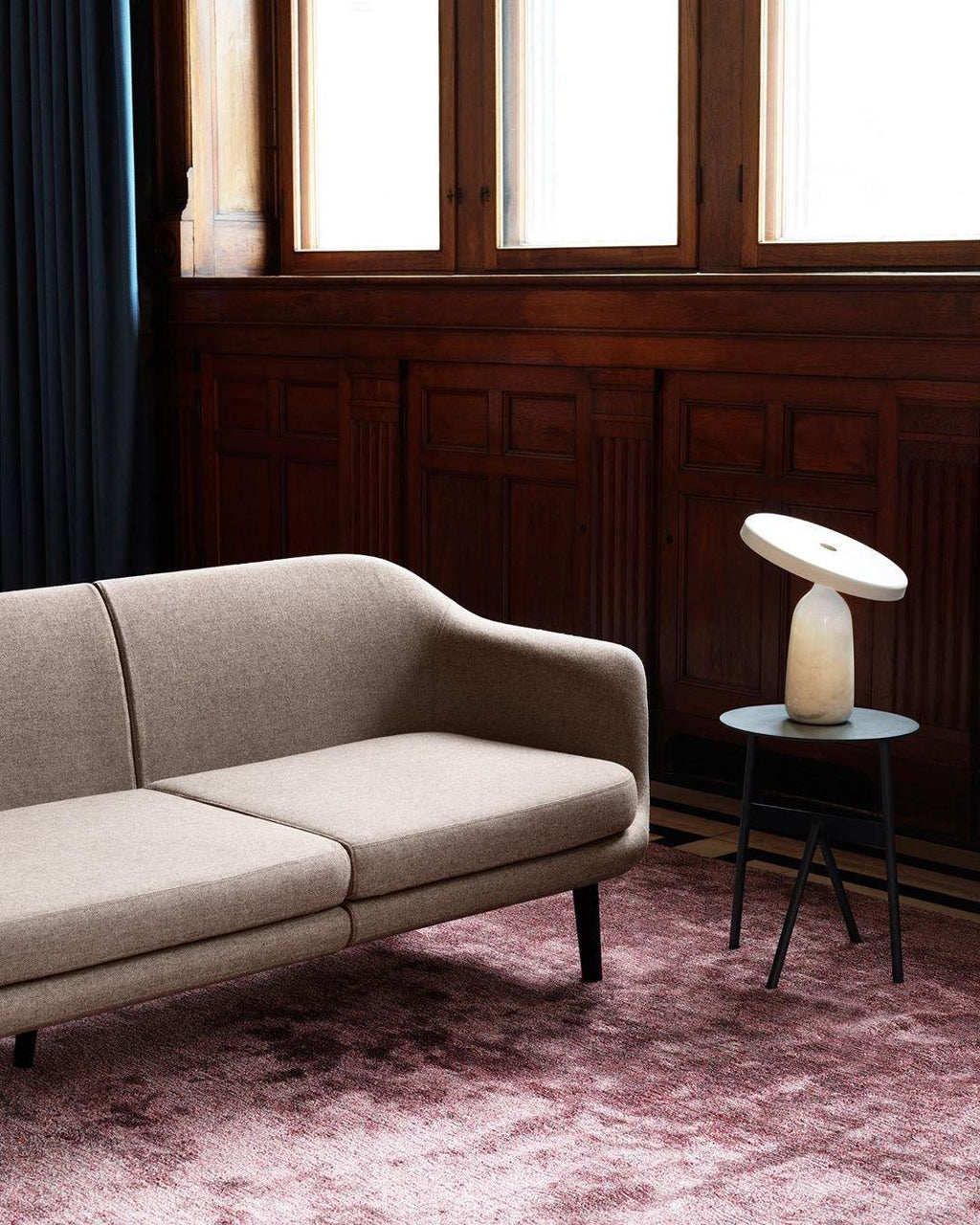 White marble Clérachic table lamp on a black side table beside a beige sofa, set on a dusty pink rug in a panelled room.