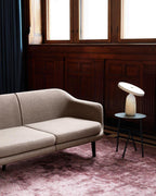 White marble Clérachic table lamp on a black side table beside a beige sofa, set on a dusty pink rug in a panelled room.