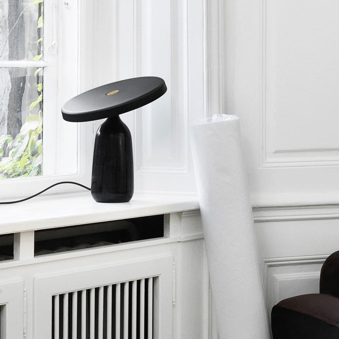 Black Clérachic table lamp on a white windowsill beside a radiator, its disc shade angled above a rolled white rug nearby.