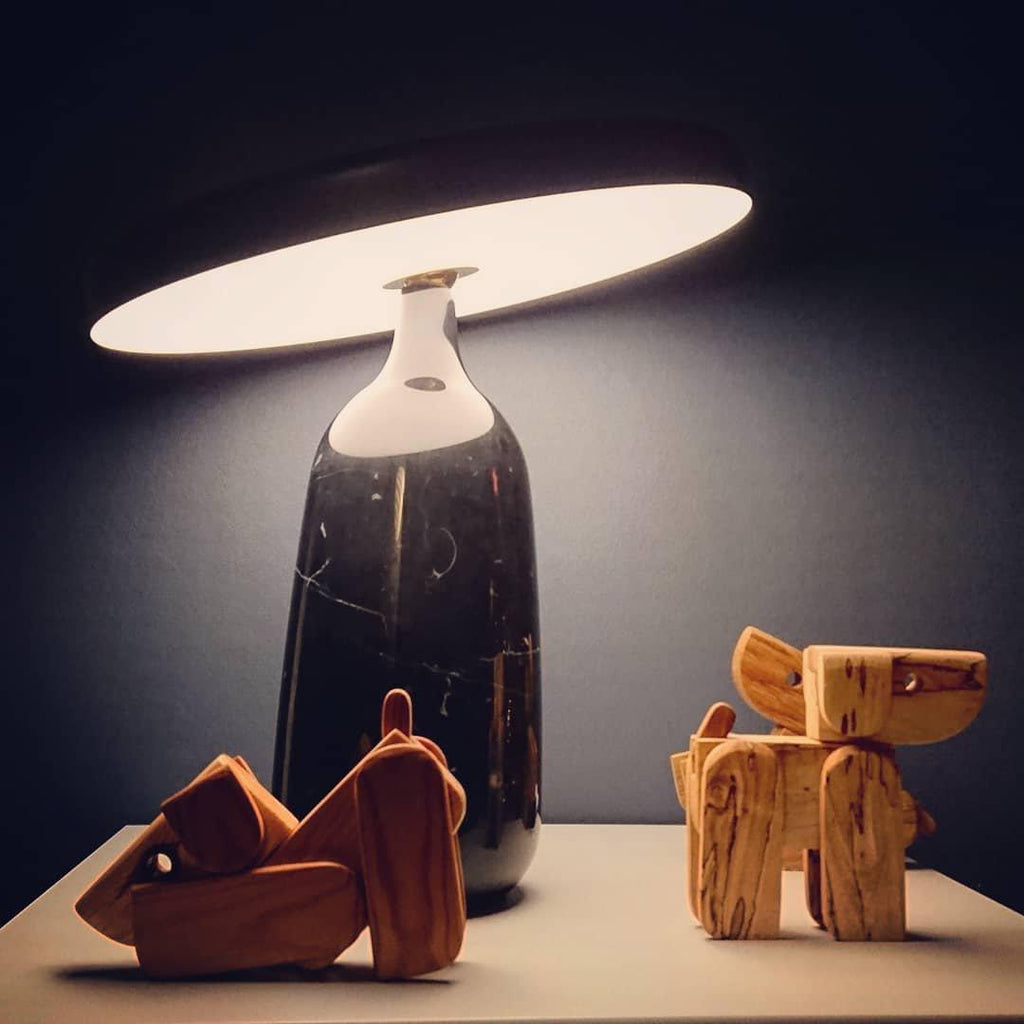 Close view of a black marble Clérachic table lamp beside wooden toy dogs, its disc shade held level in low ambient light.
