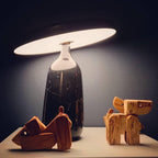 Close view of a black marble Clérachic table lamp beside wooden toy dogs, its disc shade held level in low ambient light.