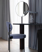Grey marble Clérachic table lamp with its disc shade tilted steeply, on a dark table beside a round mirror and a blue chair.