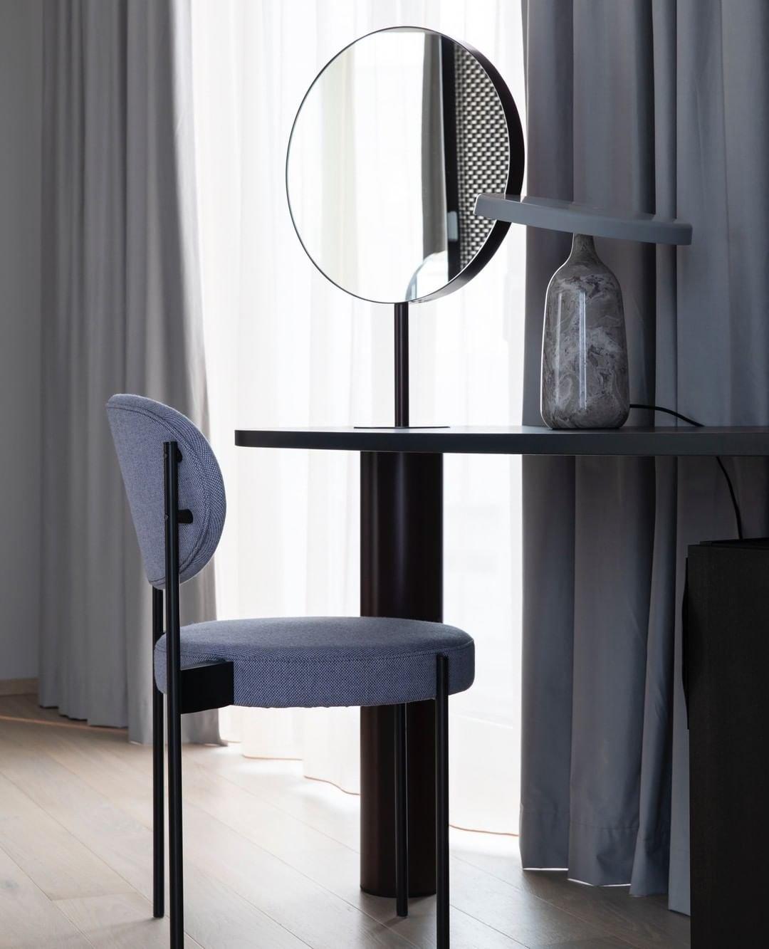 Grey marble Clérachic table lamp with its disc shade tilted steeply, on a dark table beside a round mirror and a blue chair.