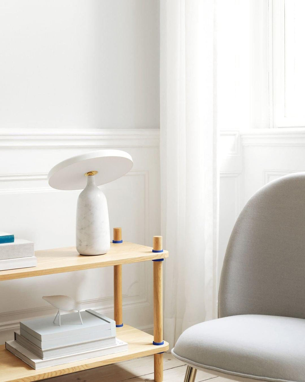 White marble Clérachic table lamp on a lower wooden shelf tier, blue-covered books stacked beside a grey armchair.