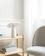 White marble Clérachic table lamp on a lower wooden shelf tier, blue-covered books stacked beside a grey armchair.