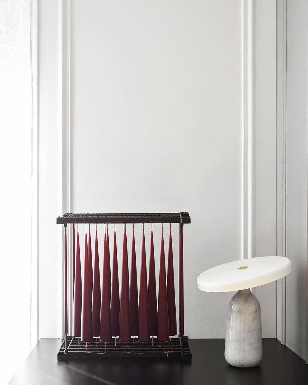 White marble Clérachic table lamp beside a dark red pleated decorative piece on a black table, against a tiled wall.