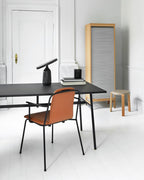 Black Clérachic table lamp lit on a black desk in a white study, its disc shade glowing beside an orange chair.