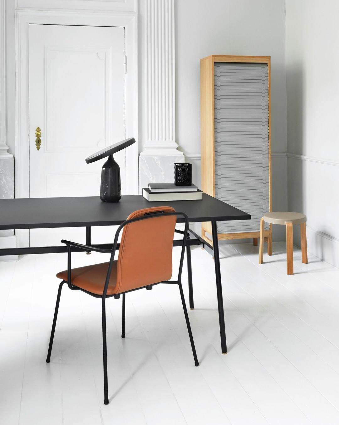 Black Clérachic table lamp lit on a black desk in a white study, its disc shade glowing beside an orange chair.
