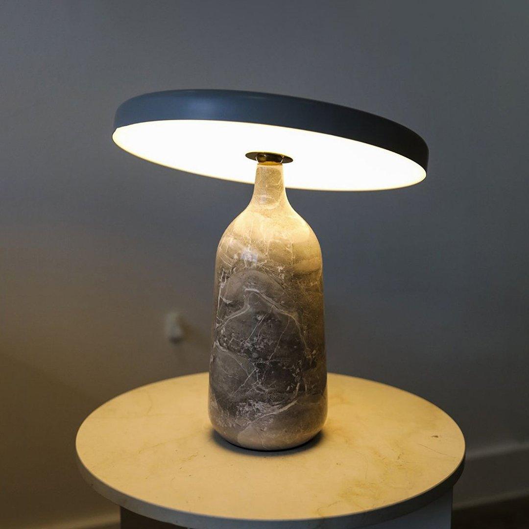 Clérachic table lamp glowing on a round wooden side table in a dim living room, its disc shade casting light downward.