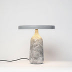 Grey marble Clérachic table lamp lit from within, its disc shade tilted slightly above the softly glowing marble base.
