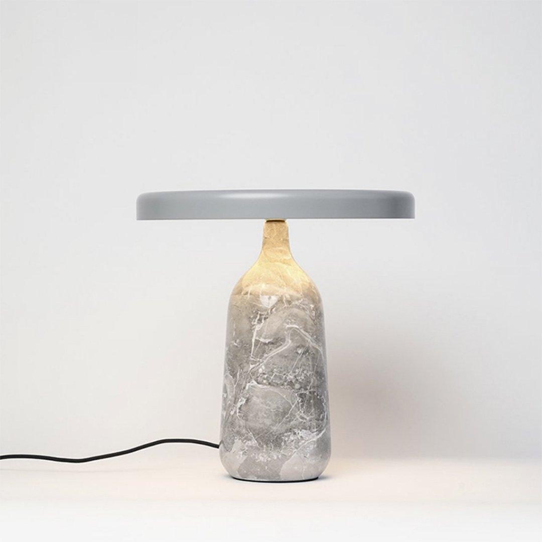 Grey marble Clérachic table lamp lit from within, its disc shade tilted slightly above the softly glowing marble base.