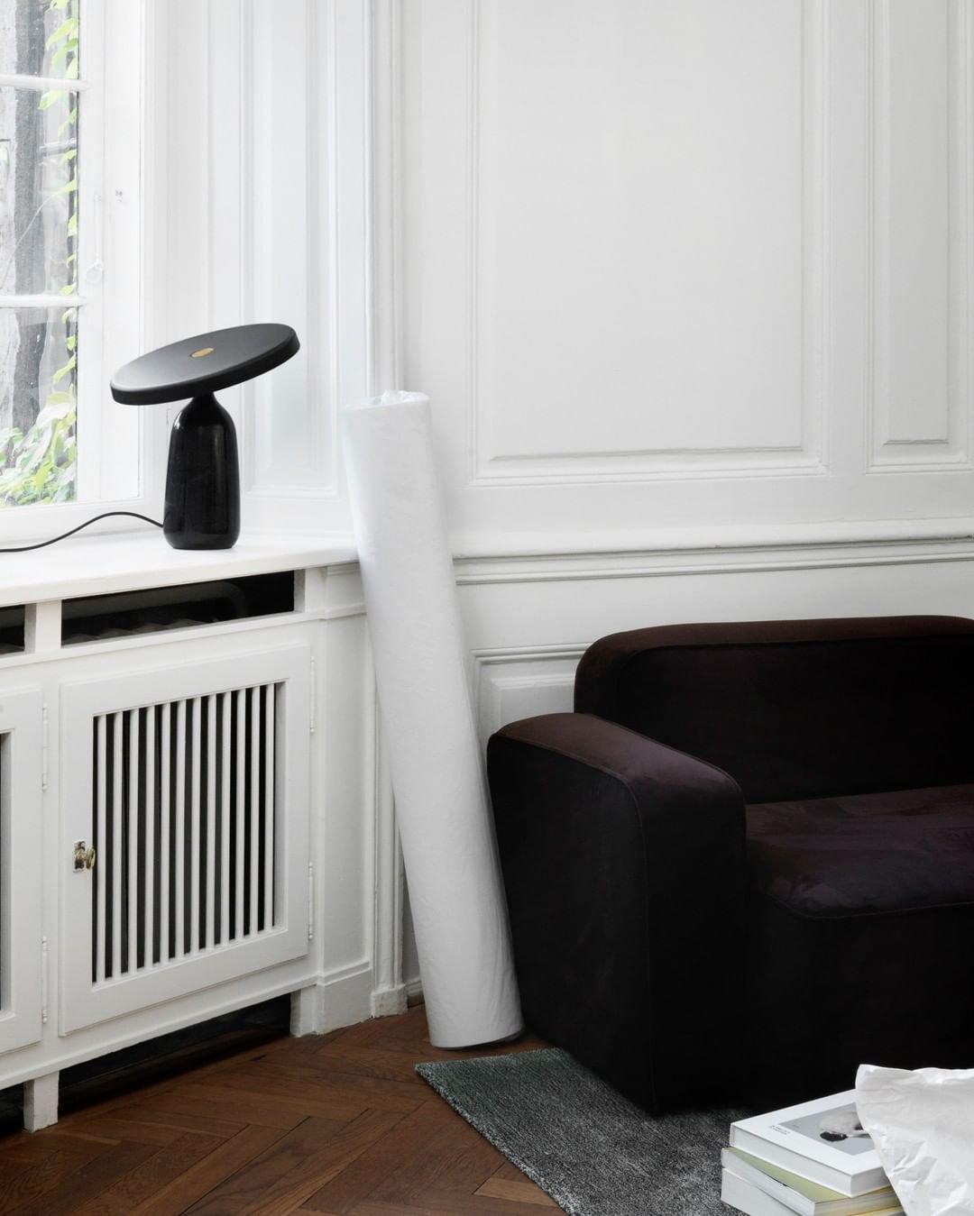 Black Clérachic table lamp on a light console table near a radiator, beside a dark brown armchair against a white wall.