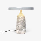 Grey marble Clérachic table lamp lit warm against a plain background, its disc shade held level above the softly glowing base.