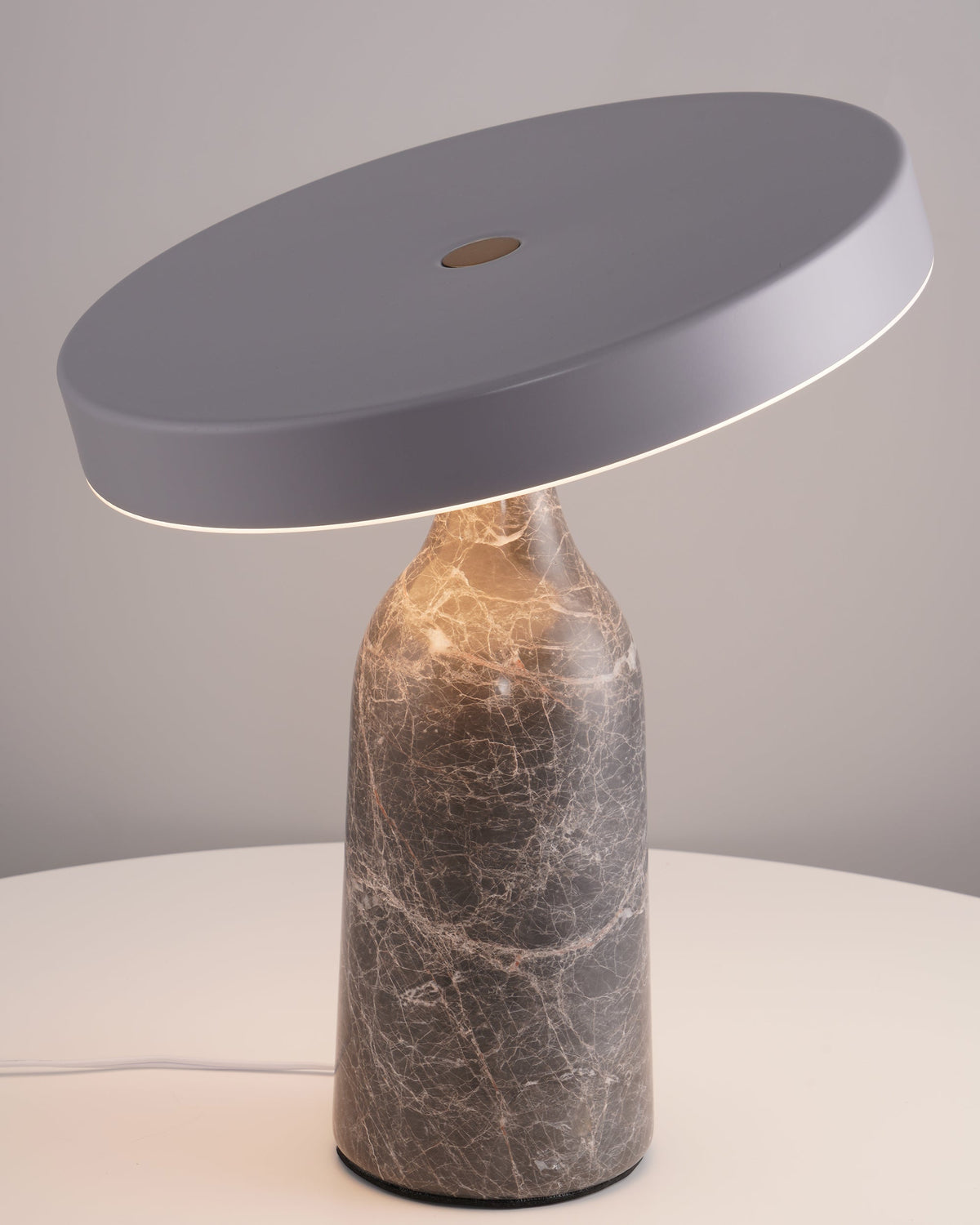 Grey marble Clérachic table lamp lit warm on a round white side table, its disc shade tilted above the glow.