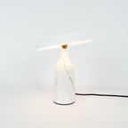 White marble Clérachic table lamp with its disc shade tilted at a steep angle, shown with its cord on white.