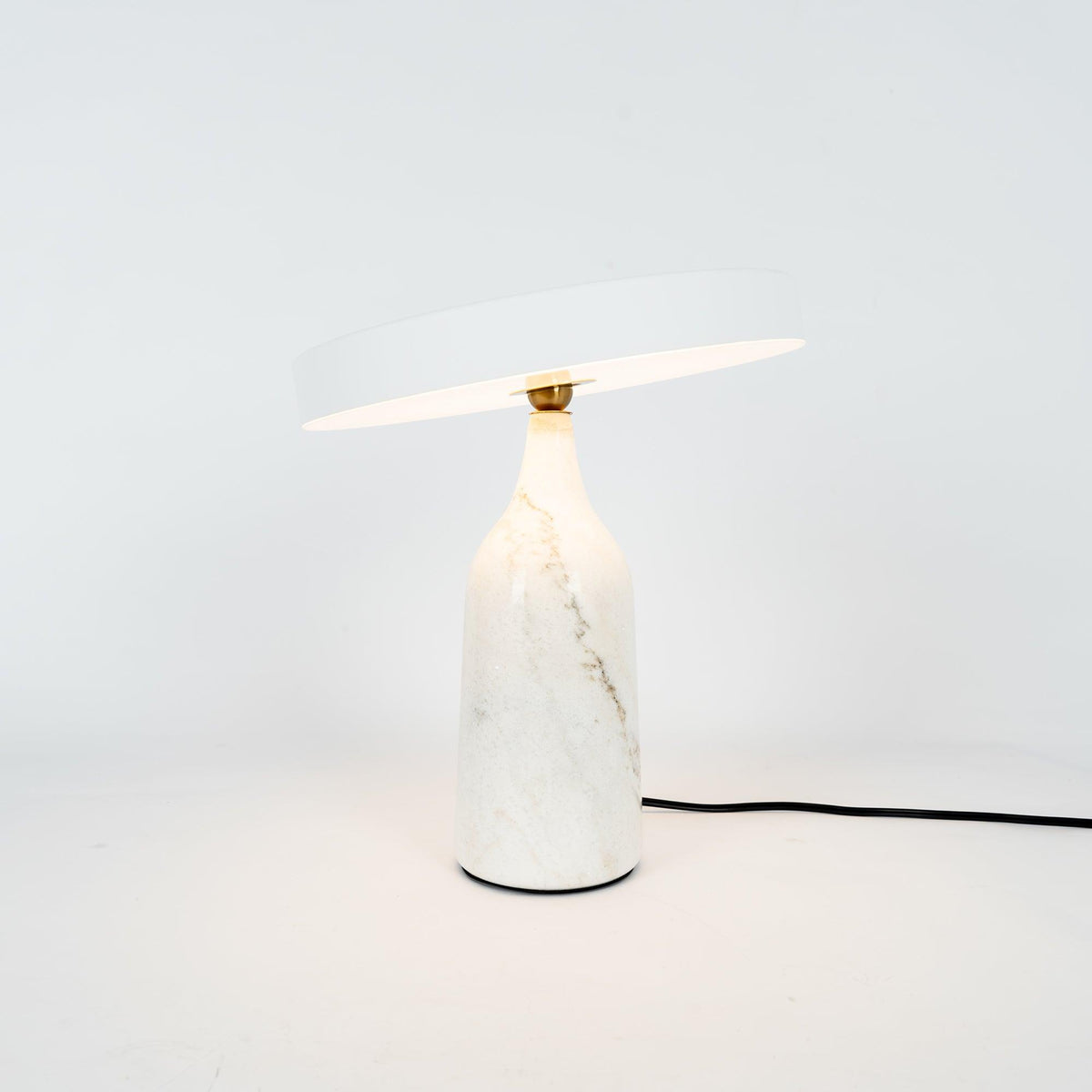 White marble Clérachic table lamp with its disc shade tilted at a steep angle, shown with its cord on white.