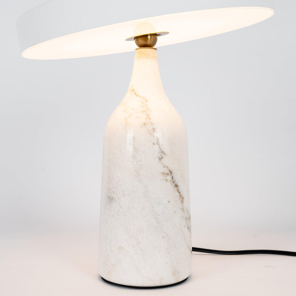 White marble Clérachic table lamp lit warm, its disc shade tilted above the glowing base, in a tall studio crop.