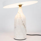 White marble Clérachic table lamp lit warm, its disc shade tilted above the glowing base, in a tall studio crop.