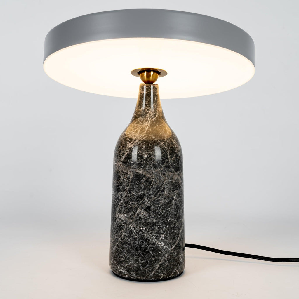 Grey marble Clérachic table lamp lit warm, its disc shade level above the glowing base on a plain background.