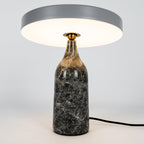 Grey marble Clérachic table lamp lit warm, its disc shade level above the glowing base on a plain background.