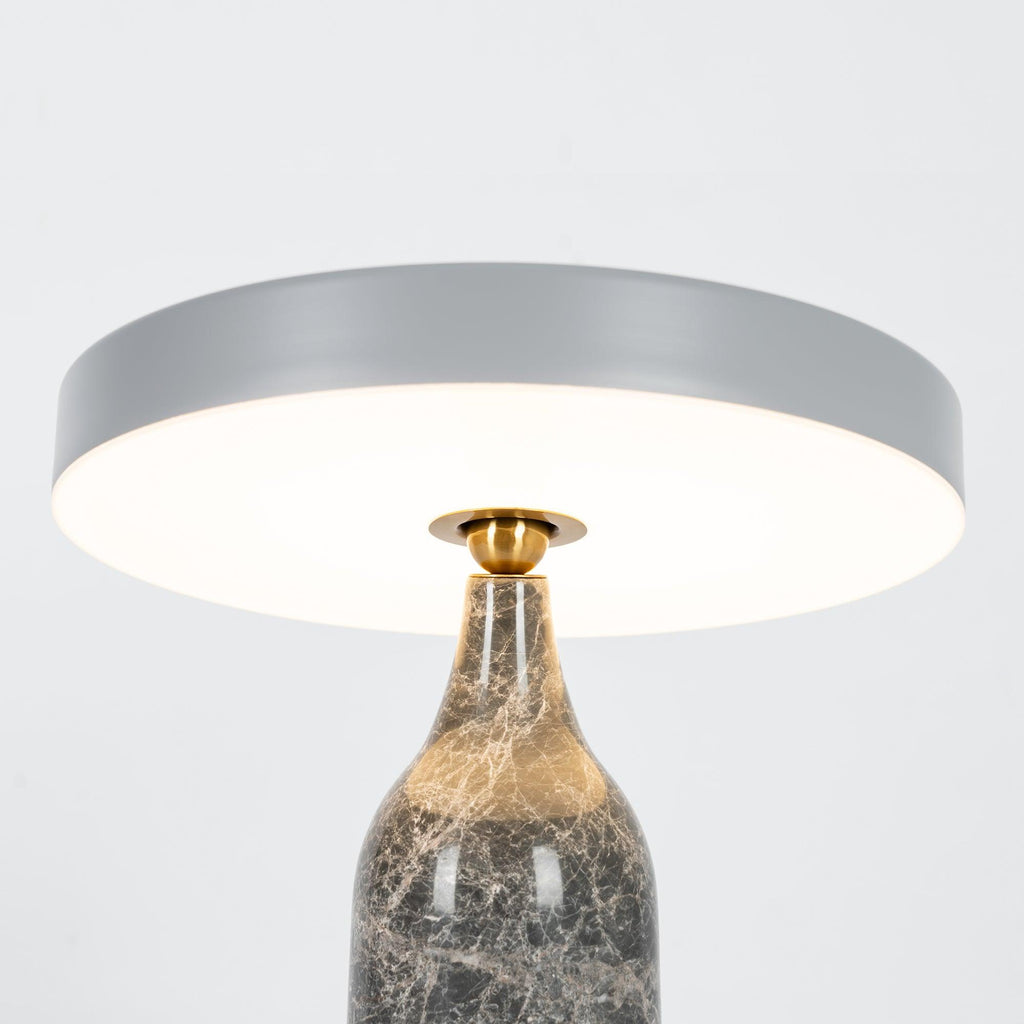 Close crop of the lit Clérachic table lamp, its grey disc shade glowing warm above the marble neck.